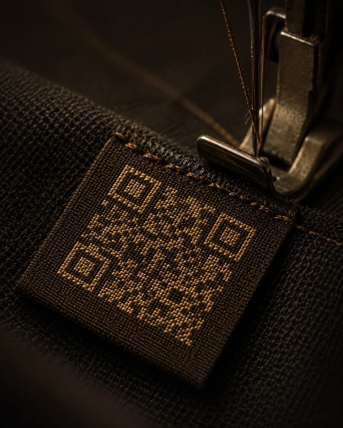 Close-up of a woven QR-code label in gold thread being stitched onto dark fabric under a sewing machine